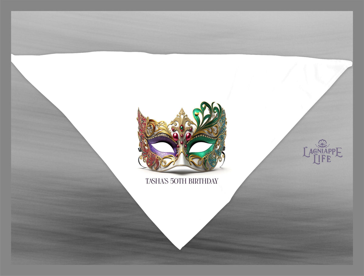 May include: A white triangular bandana with a gold, green, purple, and red masquerade mask in the center. The text 'TASHAS 50TH BIRTHDAY' is below the mask. The text 'LAGNIAPPE LIFE' is in the bottom right corner of the bandana.