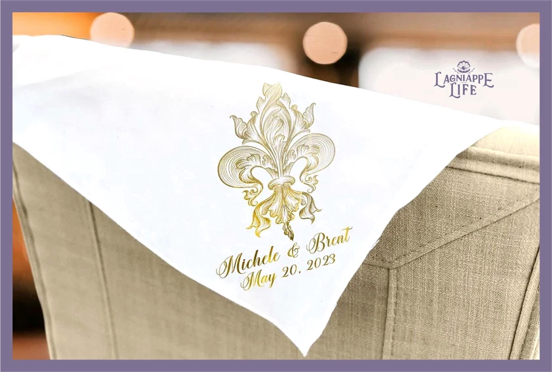 May include: White linen napkin with a gold fleur-de-lis design and the text &#39;Michele & Brent May 20, 2023&#39;.
