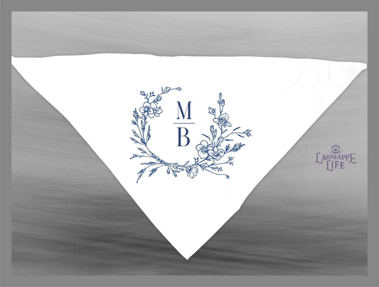 May include: A white triangle shaped napkin with a blue floral wreath design and the letters &#39;M&#39; and &#39;B&#39; in the center. The letters are separated by a horizontal line. The text &#39;LAGNIAPPE LIFE&#39; is in the lower right corner of the napkin.